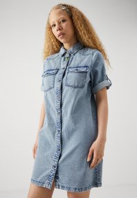 Šviesiai mėlyna džinsinė suknelė, turinti sagų užsegimą, apykaklę, dvi krūtinės kišenes ir susuktas rankoves su kontrastingu siuvinėjimu.