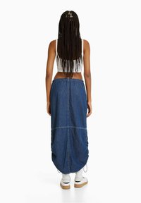 Falda maxi de denim con dobladillo fruncido, presenta un detalle de cordón y costuras en contraste. Combinada con zapatillas blancas y calcetines hasta el tobillo.