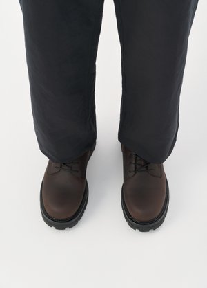 Braune Leder-Schnürschuhe mit runder Zehenform, schwarzer Gummisohle und strukturierten Details, kombiniert mit locker sitzenden dunklen Hosen.