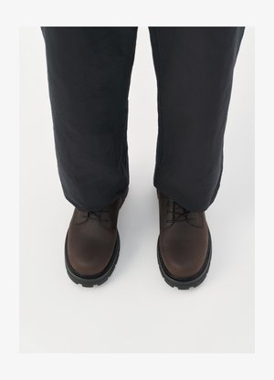 Scarpe stringate in pelle marrone con punta arrotondata, suola in gomma nera e dettagli testurizzati, abbinate a pantaloni larghi scuri.