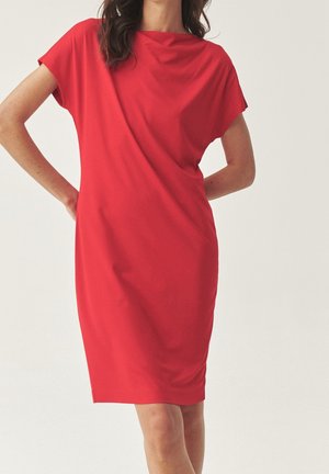 Mujer con vestido rojo brillante de manga corta hasta la rodilla, de corte holgado, de pie con las manos en las caderas sobre un fondo claro y liso.