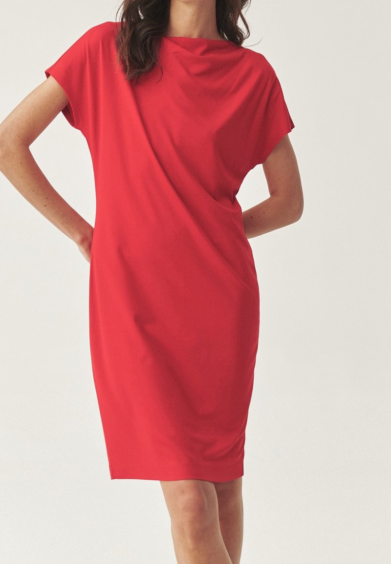 Femme portant une robe rouge vif à manches courtes et longueur genou, coupe décontractée, debout, les mains sur les hanches, sur un fond clair uni.