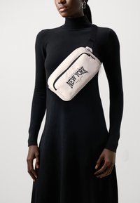 Bolsa crossbody em nylon rosa claro com um bolso frontal com fecho e "NEW YORK" impresso em letras grandes pretas, usada sobre um vestido longo preto de mangas.