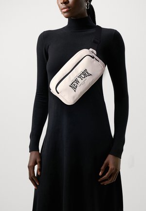 Sac à bandoulière en nylon rose clair avec une poche zippée à l'avant et "NEW YORK" imprimé en lettres noires en gras, porté sur une robe noire à manches longues.