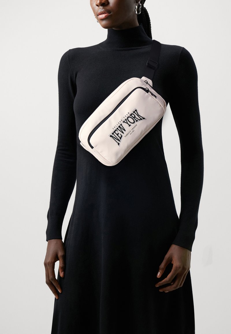 Bolsa crossbody em nylon rosa claro com um bolso frontal com fecho e "NEW YORK" impresso em letras grandes pretas, usada sobre um vestido longo preto de mangas.