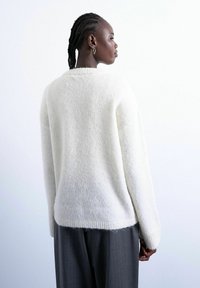 Femme noire aux cheveux tressés portant un pull blanc en laine et un pantalon gris, vue de profil sur un fond blanc uni.