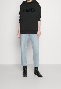 Felpa nera con cappuccio, tasca frontale e logo in peluche, abbinata a jeans azzurri a gamba dritta e stivali neri alla caviglia. Tessuto morbido.