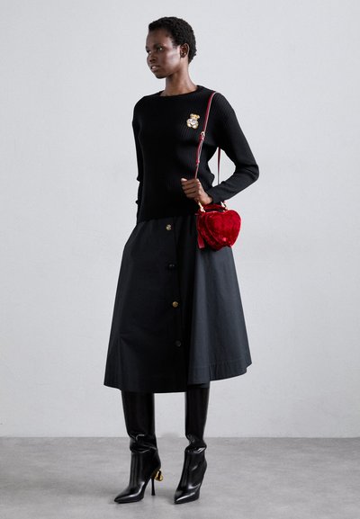 Pull noir à côtes avec une broche en forme d'ours, associé à une jupe sombre ornée de boutons dorés et des bottes noires à talons aiguilles montantes. Sac en forme de cœur rouge.