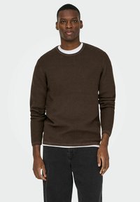 Pull long en tricot marron texturé avec un col rond, superposé sur une chemise blanche, associé à un jean en denim foncé.