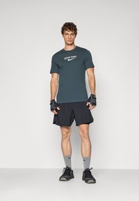 Tamsiai mėlynos Nike Pro marškinėliai vyrams su balta logotipu, derinami su juodomis šortais, pilkais kojinėmis ir juodais treniruočių batais. Trumpomis rankovėmis ir prigludusio dizaino.