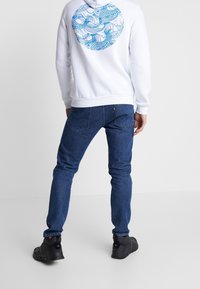 Felpa bianca con motivo a onde blu sulla schiena, abbinata a jeans blu scuri e scarpe nere. Fit rilassato e design casual.