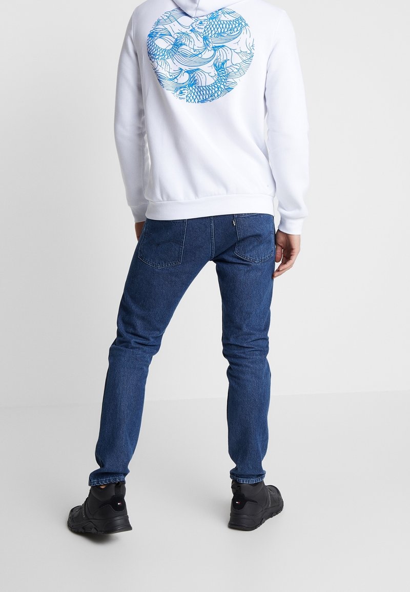 Felpa bianca con motivo a onde blu sulla schiena, abbinata a jeans blu scuri e scarpe nere. Fit rilassato e design casual.