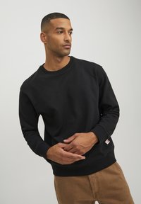 Schwarzer Baumwollpullover mit rundem Halsausschnitt und langen Ärmeln. Mit gerippten Bündchen und Saum sowie einem kleinen Logotag am unteren Ende.