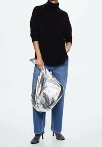 Maglione a collo alto nero, jeans a vita alta blu, mentre tiene una borsa argentata lucida. Tacchi stiletto neri e una superficie liscia e testurizzata sulla borsa.