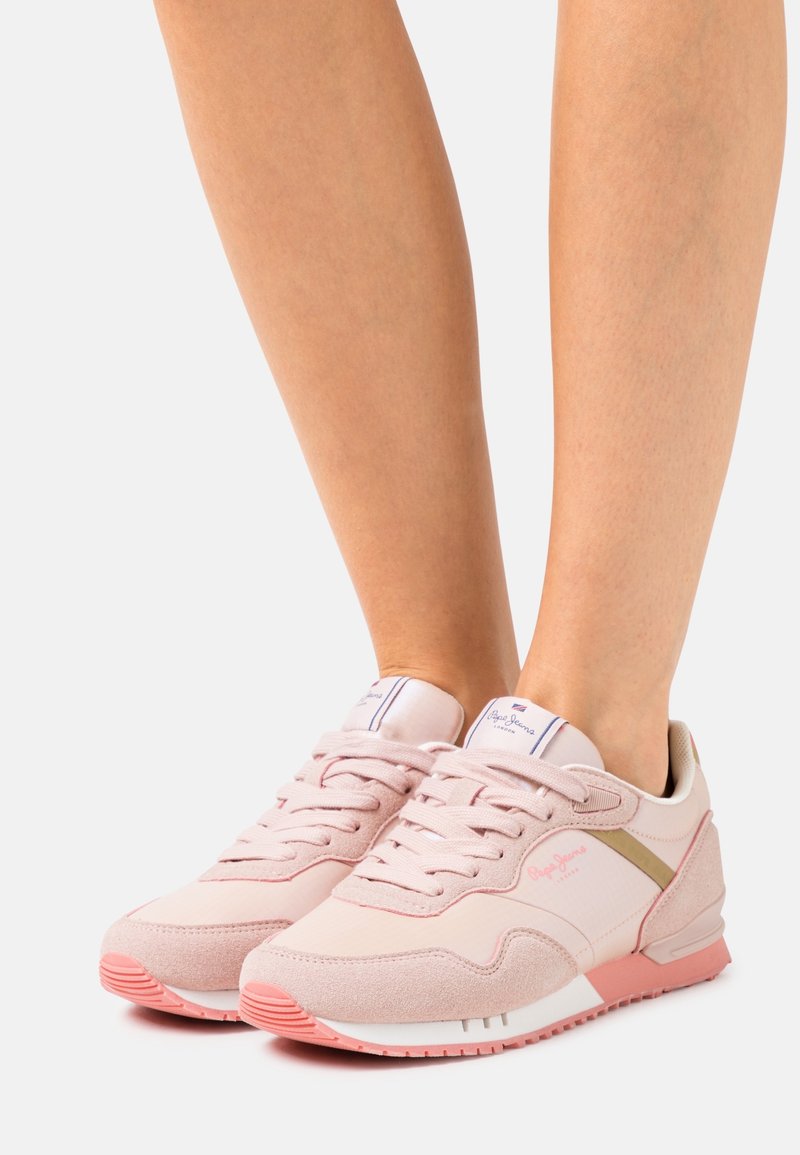 Baskets à lacets rose clair avec semelles blanches et marquage subtil, portées sur des jambes nues contre un fond blanc uni.