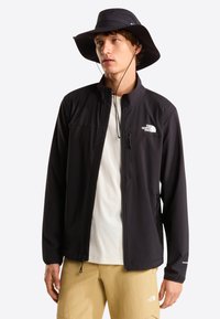 Μαύρο σακάκι softshell με φερμουάρ μπροστά, ψηλό γιακά και λογότυπο. Συνδυάζεται με λευκό πουκάμισο και μπεζ σορτς, κάτω από ένα καπέλο με φαρδύ γείσο.