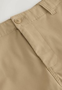 Gros plan sur la ceinture d'un pantalon beige avec fermeture par bouton et couture visible autour de la passants de ceinture et de la zone de la braguette.