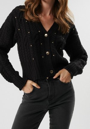Femme portant un cardigan noir en maille torsadée avec des boutons dorés et des petits clous, associé à un jean noir taille haute, mains dans les poches.