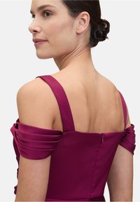 Dopasowana sukienka w głębokim kolorze magenta, z odkrytymi ramionami, plisowanymi ramiączkami i dyskretnym zamkiem z tyłu, prezentująca gładką fakturę.