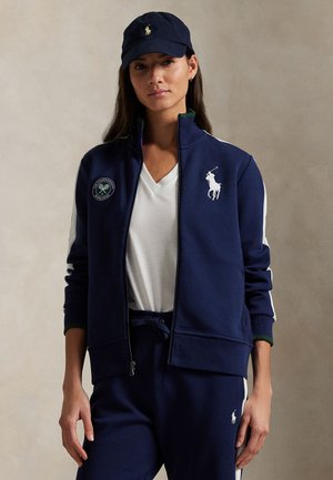 Marineblaue Reißverschlussjacke mit weißen Streifen, ausgestattet mit einem Wimbledon-Patch und einem bestickten Logo. Getragen mit einem weißen V-Ausschnitt-Shirt und passenden Hosen.
