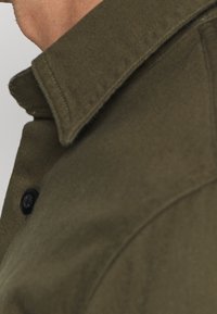 Olivgrünes Hemd mit Kragen und strukturierter Stoffqualität; verfügt über eine Knopfleiste und einen sauberen, strukturierten Kragen.