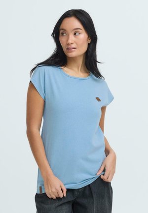 Femme aux longs cheveux noirs portant une chemise bleu clair à manches courtes et un pantalon sombre, regardant vers sa gauche avec les mains près de la taille.