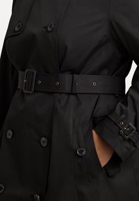 Svart trenchcoat med dubbelknäppta knappar, ett bälte med en guldfärgad spänne och textil med struktur. Har sidofickor och justerbara ärmsnoddar.