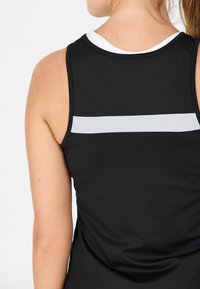 Donna che indossa un top atletico senza maniche nero con una striscia riflettente orizzontale sulla parte superiore della schiena sopra una maglietta bianca.