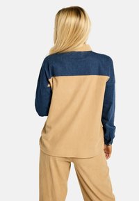 Két tónusú ing, barna és kék színben, bordázott textúrával. Hosszú ujjú, gombos mandzsettával és galléros nyakkal. Egy pár hozzáillő barna kordbársony nadrág.