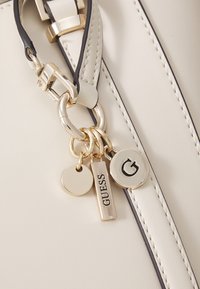 Borsa a mano in pelle crema con dettagli neri; presenta hardware dorato e charms decorativi tra cui un’etichetta rotonda e un’etichetta rettangolare "GUESS".
