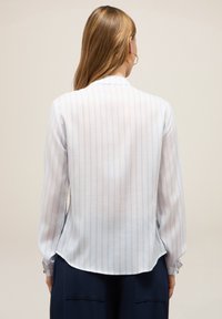 Blusa leggera e trasparente con strisce verticali blu su sfondo bianco. Presenta maniche lunghe e un orlo curvato.