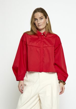 Naine, kes kannab punast pika varrukaga pluusi suurte varrukatega ja valgeid kõrge vöökohaga laia säärega pükse, seistes ühevärvilise valge tausta ees.