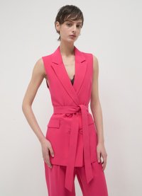 Blazer sans manches rose vif avec un col en V profond, une taille ceinturée et des accents à deux boutons, associé à un pantalon large assorti.