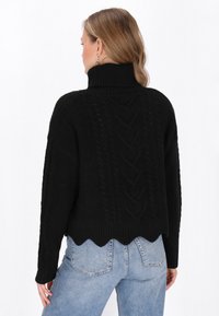 Schwarzer Strickpullover mit hohem, geripptem Kragen, Kabelstrick-Details, cropped Länge und gewelltem Saum; getragen mit hellblauen Jeans.