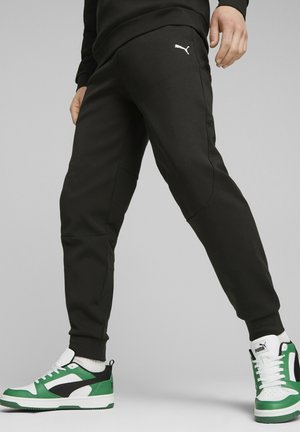Pantalones negros ajustados hechos de una tela suave, con un logo blanco. Combinados con zapatillas verdes y blancas, que muestran un diseño deportivo.