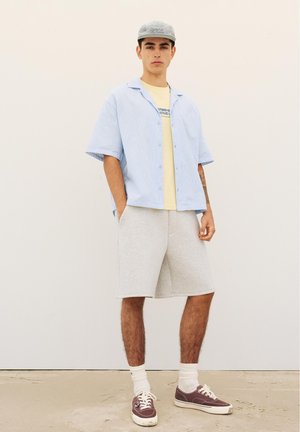 Mladý muž nosi svetlomodrú pruhovanú overshirt, žlté grafické tričko, sivé šortky, biele ponožky, fialové tenisky a sivú čiapku, stojí s rukami v vreckách.