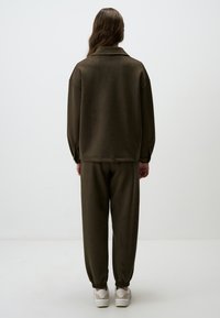 Completo da tuta verde scuro, morbido e dalla vestibilità rilassata, con colletto, polsini e orlo elasticizzati. Abbinato a sneakers bianche.
