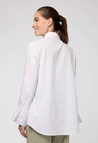 Camicia bianca a maniche lunghe con una vestibilità comoda, caratterizzata da un colletto alto e polsini spaccati, realizzata in tessuto morbido, indossata sopra pantaloni di colore chiaro.