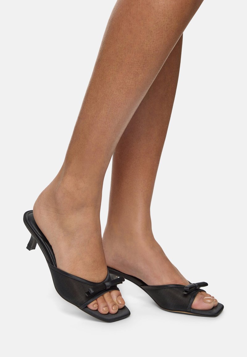 Mules noires à talon avec un bout carré, dotées d'un dessus en tissu, d'un accent en nœud et d'un petit talon kitten. Texture lisse sur l'ensemble.