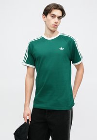 Πράσινο βαμβακερό μπλουζάκι με λευκές λεπτομέρειες, διαθέτει στρογγυλή λαιμόκοψη και τρεις λευκές ρίγες σε κάθε ώμο. Λογότυπο Adidas σε λευκό στο στήθος.