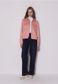 Vrouw die een roze jas, een witte coltrui, donkere spijkerbroek en witte sneakers draagt, staand tegen een effen witte achtergrond.