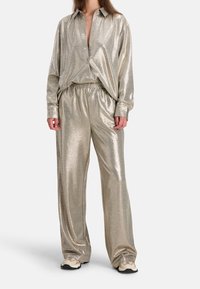 Das metallic goldene Loungewear-Set umfasst ein lockeres Hemd mit Knöpfen und Pants mit elastischem Bund, die eine glatte, glänzende Textur mit dezenten Mustern aufweisen.