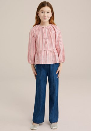 Blusa a righe rossa e bianca con maniche lunghe, scollo arricciato e dettagli con bottoni, abbinata a pantaloni in denim a gamba larga blu e scarpe chiare.