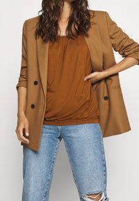 Blazer marrone sopra una maglia ruggine, abbinato a jeans chiari strappati. Il blazer ha due bottoni e maniche arrotolate. Tessuto dalla texture liscia.