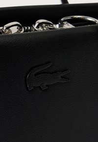 Μαύρη τσάντα από δέρμα με υφή, με ανάγλυφο λογότυπο κροκόδειλου και ασημένια αλυσίδα ως μεταλλικό στοιχείο εμφανές μέσα στην τσάντα.