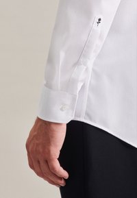 Chemise blanche avec poignets boutonnés, ornée d'un motif floral noir brodé. Tissu lisse et coupe ajustée.