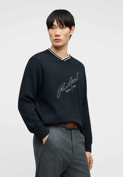 Νέος άνδρας με μαύρα μαλλιά που φοράει μαύρο φούτερ με λευκή γραφή και γκρι παντελόνι, όρθιος με το ένα χέρι στην τσέπη.