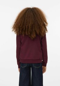 Burgunderry sweatshirt med ribbade manschetter och nederkant, bakvy visar en texturerad, voluminös lockig frisyr och denimbyxor med vida ben.