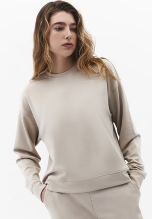 Frau mit langen, welligen Haaren, die einen beigen Pullover und passende Hosen trägt, Hände in den Taschen, schaut leicht zur Seite.