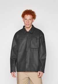 Veste noire texturée à manches longues, col et poche poitrine. Le modèle porte un pantalon de couleur claire. Design simple et coupe décontractée.